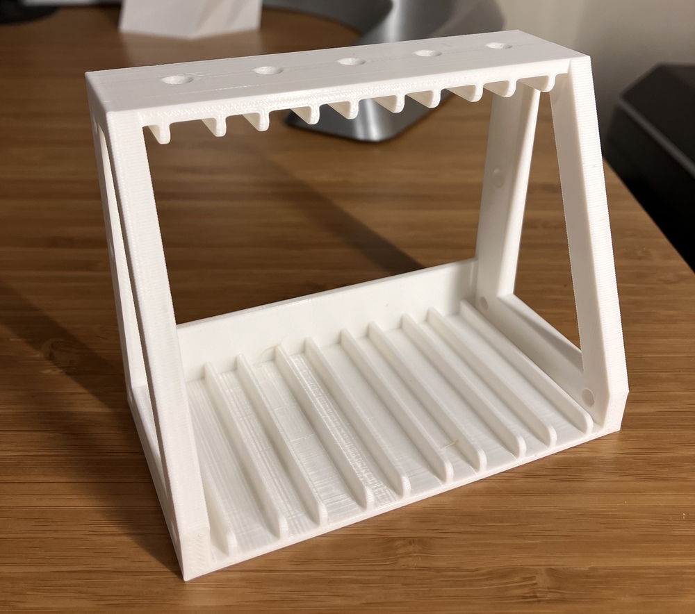 A white 3D printed part. The left and right sides are triangles while the top and bottom are rectangles. 5 slots are visible on the inside bottom of the part. On the top are 5 countersunk holes for securing the SSDs in place with a screw.