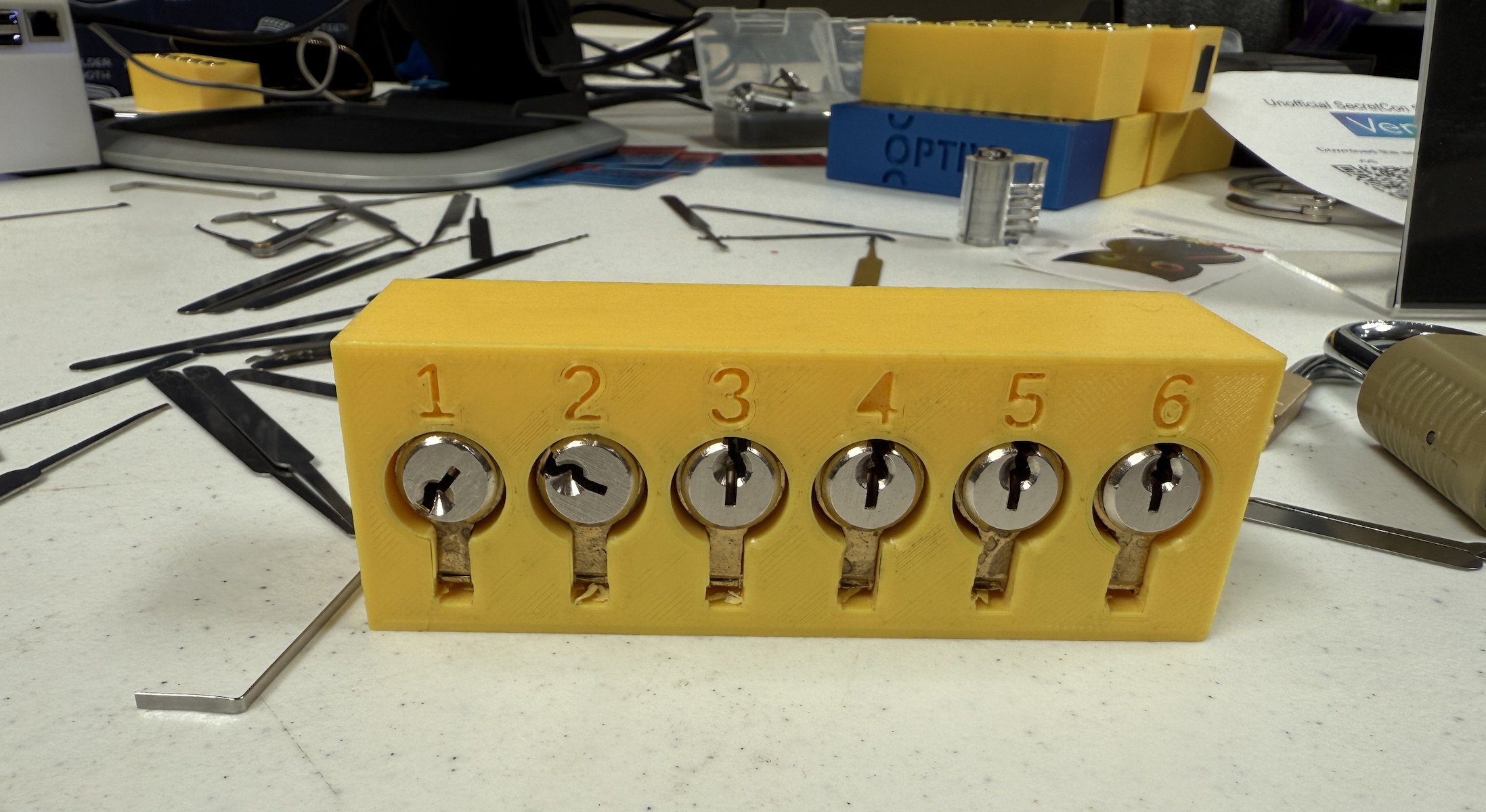Close up on a table covered in lock picks, tension wrenches, and in the foreground a yellow plastic rectangle with 6 circular lock faces embedded.